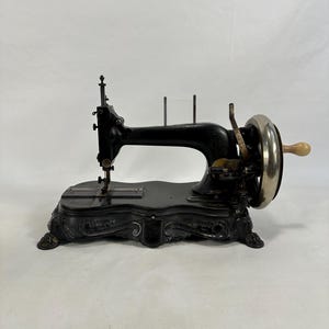 May include: An antique black sewing machine with a decorative base and a wooden hand crank. The machine has a metal needle plate and a foot pedal.