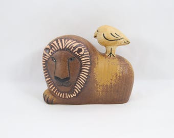 Puede incluir: Escultura de cerámica de un león con un pájaro posado en su espalda. El león es marrón con una melena y rasgos faciales detallados. El pájaro es amarillo con detalles negros. Fondo blanco.