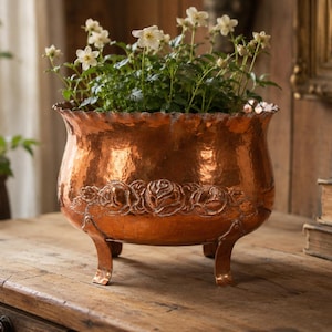 May include: A hammered copper planter with a scalloped rim and three curved legs. The planter features a decorative band of embossed roses and is filled with green plants and white flowers. It sits on a wooden table.