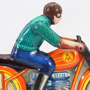 May include: Vintage toy motorcycle with a rider. The motorcycle is primarily orange with yellow and red accents. The rider wears a brown helmet, a green shirt, and blue pants. The toy has a classic, retro design.