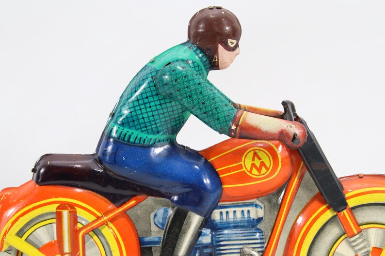 May include: Vintage toy motorcycle with a rider. The motorcycle is primarily orange with yellow and red accents. The rider wears a brown helmet, a green shirt, and blue pants. The toy has a classic, retro design.