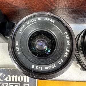 May include: A black Canon FD 28mm 1:2.8 lens with the text "LENS MADE IN JAPAN CANON LENS FD 28mm 1:2.8" printed on the side.