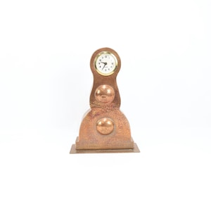 May include: A copper-colored mantel clock with a round clock face and Roman numerals. The clock has a textured surface and a decorative design with three spherical accents. The clock is approximately 10 inches tall.