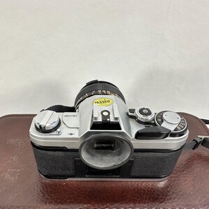 May include: A silver Canon AE-1 Program 35mm film camera with a Canon FD 50mm f/1.8 lens. The camera has a black leather grip and a black lens cap. The camera has a sticker that says "PASSED" on the lens.