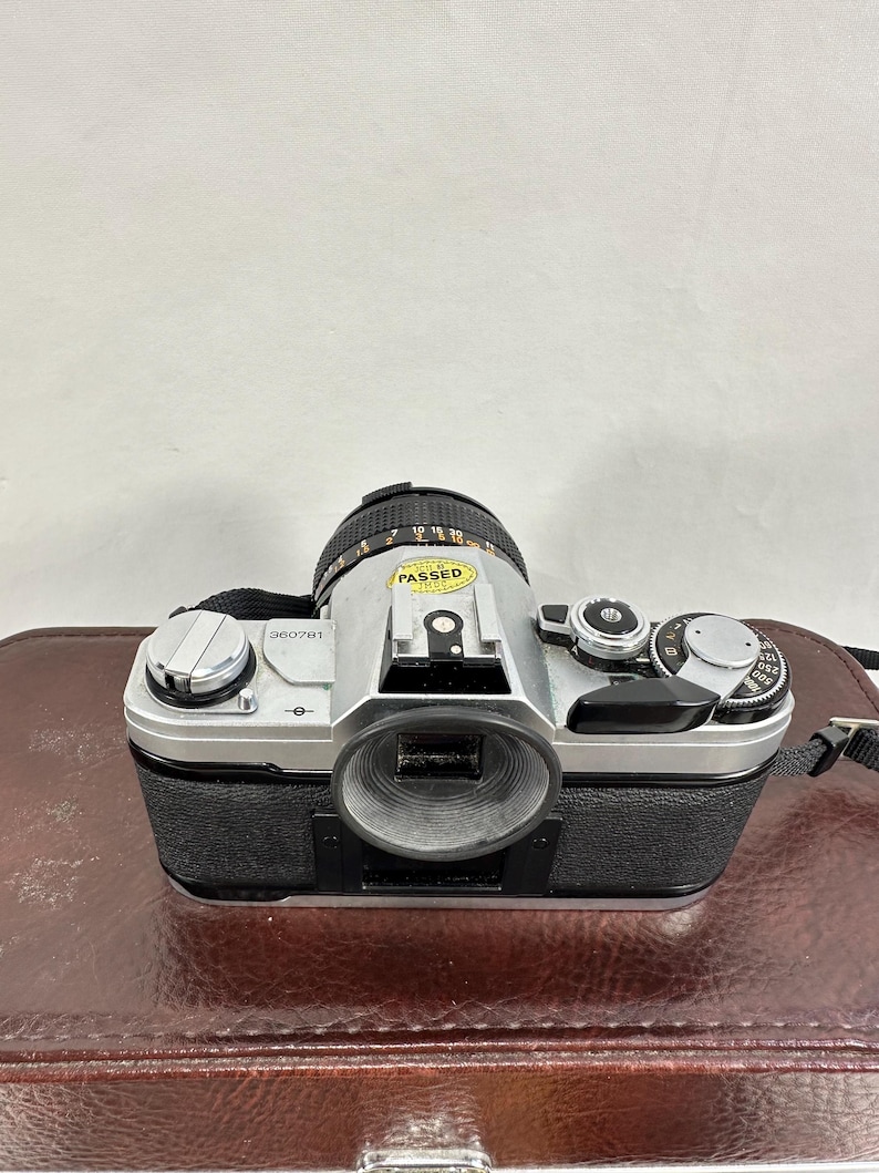 May include: A silver Canon AE-1 Program 35mm film camera with a Canon FD 50mm f/1.8 lens. The camera has a black leather grip and a black lens cap. The camera has a sticker that says "PASSED" on the lens.