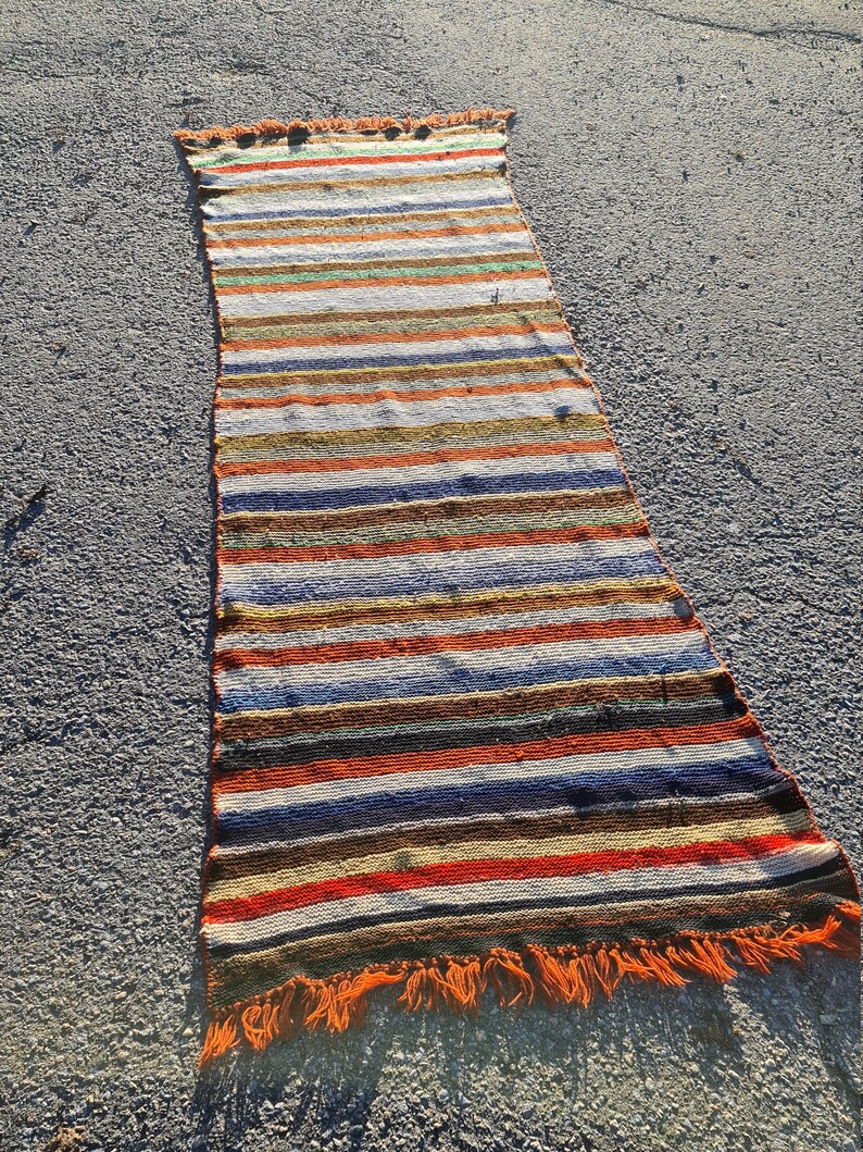 May include: A long, narrow, striped rug with orange fringe. The rug has a variety of colors, including red, orange, yellow, green, blue, and brown.
