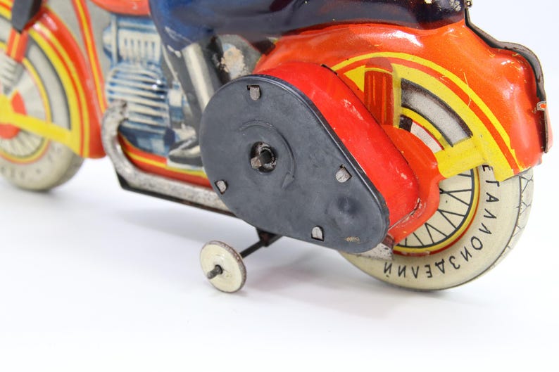 May include: Vintage toy motorcycle in red, orange, yellow, and blue. The motorcycle has a black side panel and a white tire with text. The toy has a small white wheel for balance.