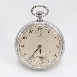 Op de afbeelding: Een vintage zilveren Omega zakhorloge met een crèmekleurige wijzerplaat. Het horloge heeft zwarte cijfers, wijzers en een subdial. Het horloge heeft een zilveren kroon en een lus voor een ketting. De wijzerplaat heeft het Omega-logo.