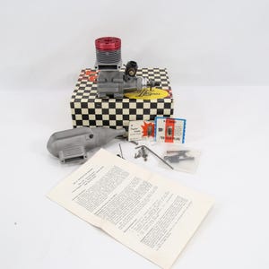 Puede incluir: Un motor de modelo con culata roja se encuentra sobre una caja a cuadros en blanco y negro. Se disponen cerca piezas de motor adicionales, bujías y un manual. El motor es plateado y negro.