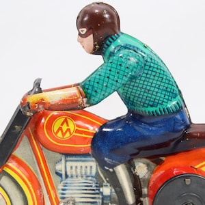 May include: Vintage toy motorcycle with a rider. The motorcycle is primarily red with yellow and black accents. The rider wears a brown helmet, a teal shirt, and blue pants. The toy has a classic, retro aesthetic.
