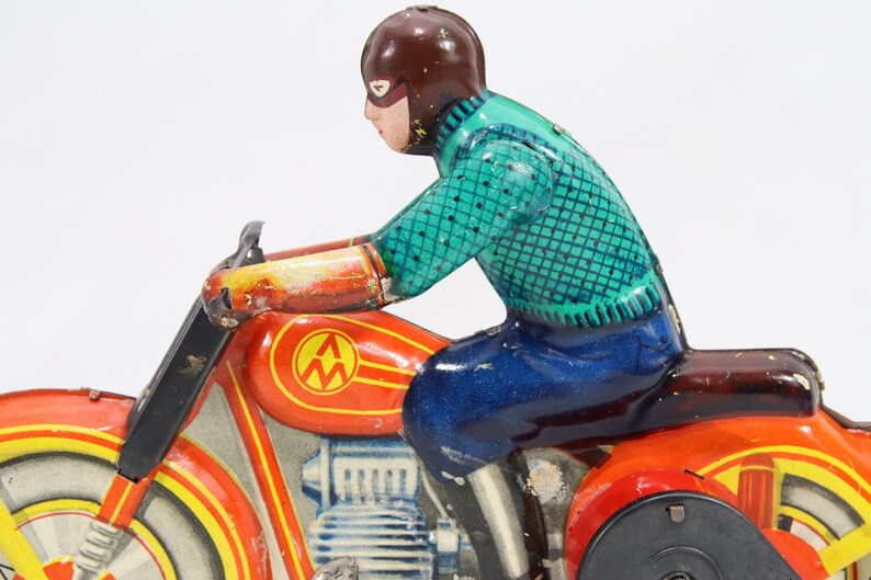 May include: Vintage toy motorcycle with a rider. The motorcycle is primarily red with yellow and black accents. The rider wears a brown helmet, a teal shirt, and blue pants. The toy has a classic, retro aesthetic.