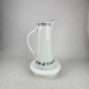 Puede incluir: Una jarra de cerámica blanca con un diseño floral azul y gris. La jarra tiene un pico y una asa. Está sentada sobre un pedestal blanco.