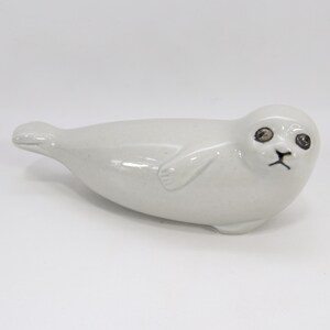 Może przedstawiać: Ceramiczna figurka foki w jasnoszarym kolorze. Foka ma czarne oczy i czarny nos. Figurka leży na białej powierzchni. Foka ma gładkie, błyszczące wykończenie. Ozdoba.