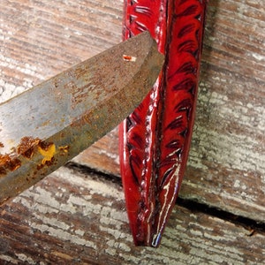 Puede incluir: Primer plano de una funda de cuero roja con un dise&ntilde;o decorativo. La funda est&aacute; parcialmente abierta, revelando una hoja de metal oxidada.