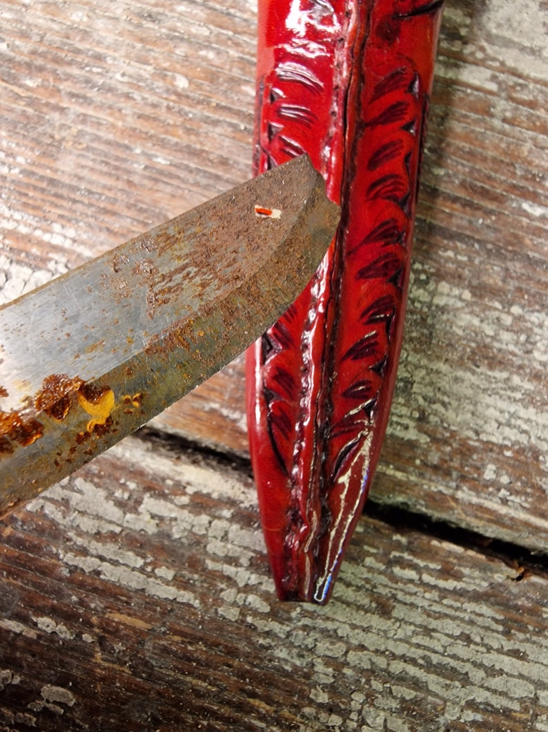 Puede incluir: Primer plano de una funda de cuero roja con un dise&ntilde;o decorativo. La funda est&aacute; parcialmente abierta, revelando una hoja de metal oxidada.