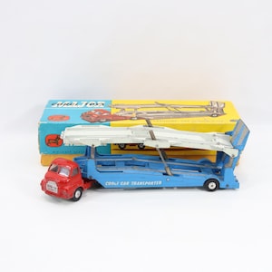 May include: Vintage toy car transporter with a red cab and a blue trailer. The trailer has a white ramp and is labeled "CORGI CAR TRANSPORTER". The box behind the toy is blue and yellow and says "CORGI TOYS".