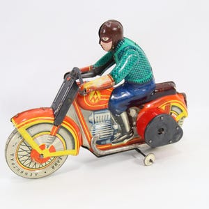 May include: Vintage toy motorcycle with a rider. The motorcycle is primarily red and orange with yellow accents. The rider wears a brown helmet, a green shirt, blue pants, and boots. The toy has a wind-up mechanism and a side wheel for balance.