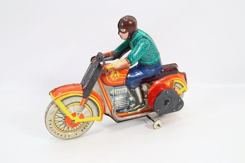 May include: Vintage toy motorcycle with a rider. The motorcycle is primarily red and orange with yellow accents. The rider wears a brown helmet, a green shirt, blue pants, and boots. The toy has a wind-up mechanism and a side wheel for balance.