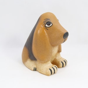 May include: A ceramic basset hound figurine in a seated position. The dog has tan fur with black accents on its ears and back. Its paws are tan with black toenails, and it has a black nose and a gentle expression.