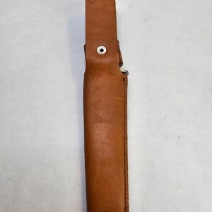 May include: A brown leather sheath with a white button and stitching. The sheath is designed to hold a knife or other sharp object.