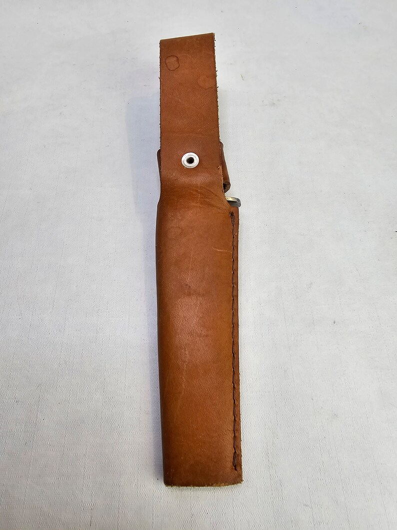 May include: A brown leather sheath with a white button and stitching. The sheath is designed to hold a knife or other sharp object.