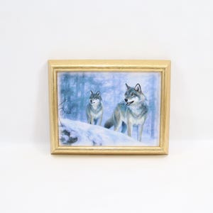 Pode incluir: Uma peça de arte de parede retangular com uma pintura de dois lobos cinzentos num cenário de floresta com neve. A obra de arte está inserida numa moldura simples de cor dourada metálica com acabamento liso e rebordo fino.