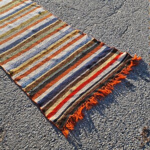 May include: A long, narrow, striped rug with orange fringe. The rug has a variety of colors, including brown, orange, white, blue, and green.