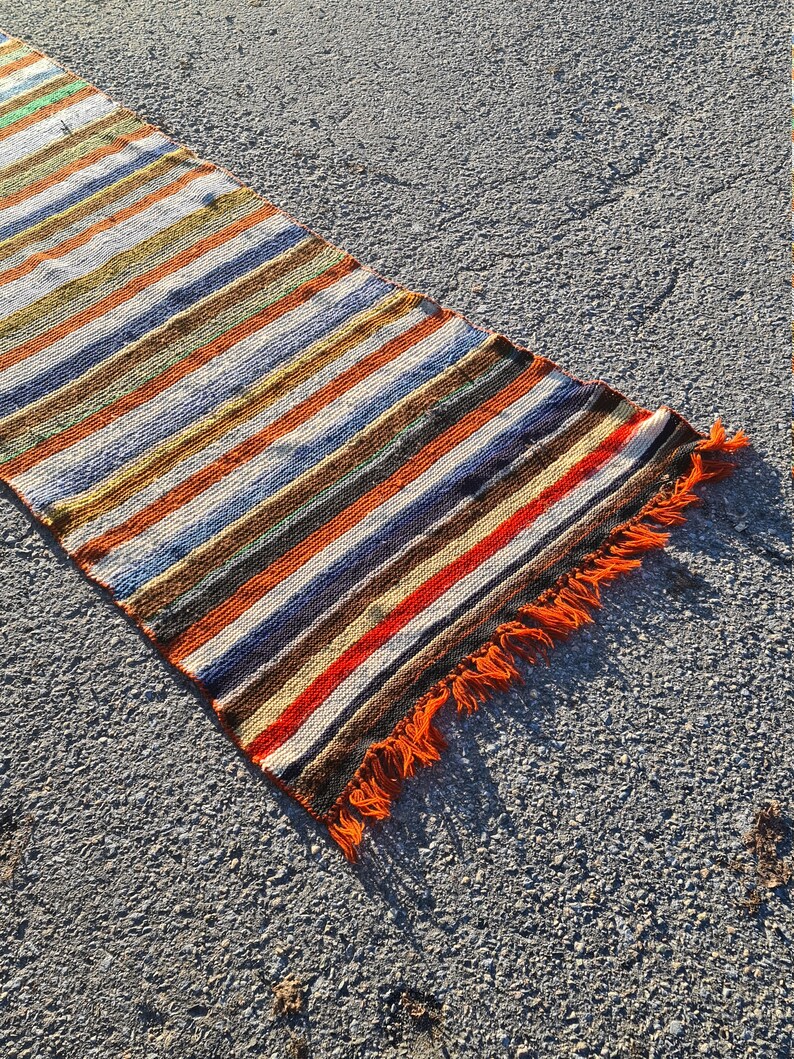 May include: A long, narrow, striped rug with orange fringe. The rug has a variety of colors, including brown, orange, white, blue, and green.
