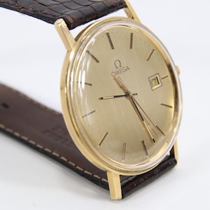 Könnte beinhalten: Eine goldfarbene Omega-Armbanduhr mit einem braunen Lederarmband. Das goldfarbene Zifferblatt hat schwarze Zeiger und Markierungen sowie ein Datumsfenster. Das Armband ist dunkelbraun mit einem strukturierten Muster.