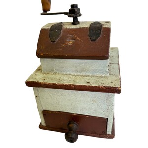 May include: Antique wooden coffee grinder with a hand crank, painted white with brown accents. The grinder has a drawer for collecting coffee grounds.