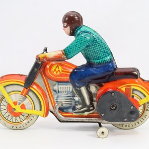 May include: Vintage toy motorcycle with a rider. The motorcycle is primarily orange and yellow with silver accents. The rider is wearing a brown helmet, a green shirt, and blue pants. The toy has a wind-up mechanism.
