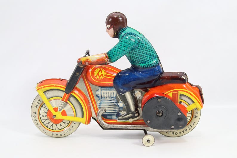 May include: Vintage toy motorcycle with a rider. The motorcycle is primarily orange and yellow with silver accents. The rider is wearing a brown helmet, a green shirt, and blue pants. The toy has a wind-up mechanism.