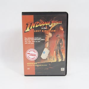 Puede incluir: Caja de videojuego negra para "Indiana Jones in the Lost Kingdom". La portada presenta el título del juego en amarillo y la silueta de un hombre. La caja tiene un fondo rojo y naranja con el texto "The ultimate challenge."