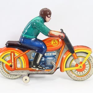 May include: Vintage toy motorcycle with a rider. The motorcycle is primarily orange with yellow and red accents, and the rider is wearing a green shirt, blue pants, and a brown helmet. The wheels have text on them.