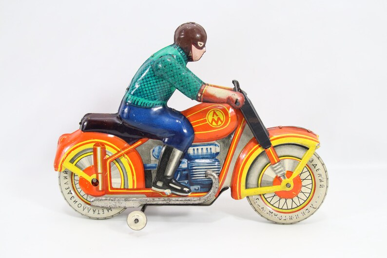 May include: Vintage toy motorcycle with a rider. The motorcycle is primarily orange with yellow and red accents, and the rider is wearing a green shirt, blue pants, and a brown helmet. The wheels have text on them.