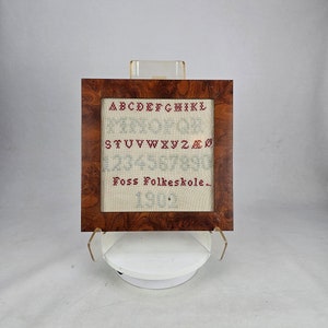 May include: A framed cross-stitch sampler featuring the alphabet in red and blue thread. The sampler also includes the text "Foss Folkeskole" and the year "1903".