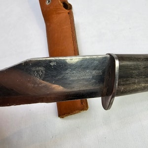 May include: A Helle-B&oslash;e "Aftenstjerne" Bowie knife with a brown leather sheath. The knife has a black wooden handle and a stainless steel blade with the text "Aftenstjerne Bowie Design S. Bergeman Made in Norway" on it.