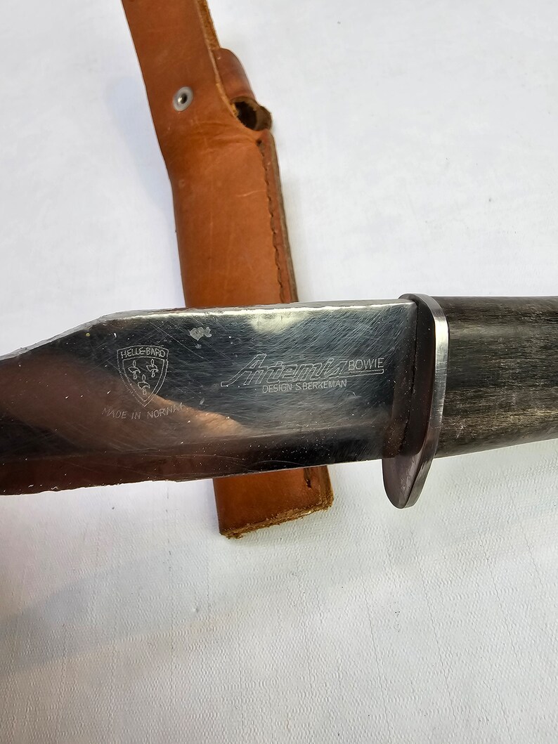 May include: A Helle-B&oslash;e "Aftenstjerne" Bowie knife with a brown leather sheath. The knife has a black wooden handle and a stainless steel blade with the text "Aftenstjerne Bowie Design S. Bergeman Made in Norway" on it.