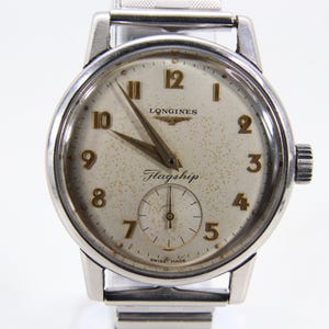 Puede incluir: Un reloj de pulsera Longines Flagship vintage con caja y correa plateadas. La esfera es blanquecina con números y agujas doradas. Una pequeña subesfera de segundos está en la parte inferior. Hecho en Suiza.