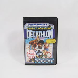 Puede incluir: Cartucho de videojuego negro Commodore 64 para Daley Thompson's Decathlon. La caja del juego presenta una colorida ilustración de un atleta compitiendo en el decatlón. El logotipo de Ocean es visible.