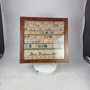 May include: A framed sampler with a wooden frame. The sampler is white with colorful embroidery of the alphabet, numbers, and the name "Ilmi Jacobsen, 1885."
