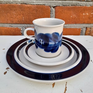 May include: A white ceramic coffee cup with a blue floral design sits on a set of three white ceramic plates with a dark blue rim.