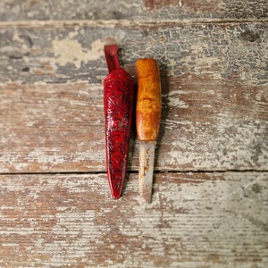 Puede incluir: Una funda de cuchillo de cuero rojo con intrincados dise&ntilde;os florales y un cuchillo con mango de madera y hoja de metal.