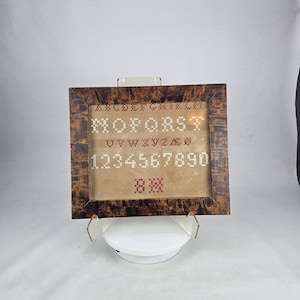May include: A framed sampler with a brown wooden frame. The sampler is made of beige fabric with red embroidery. The embroidery includes the alphabet, numbers 1 to 10, and a small cross stitch pattern.