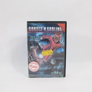 Puede incluir: Cartucho de videojuego de Ghosts 'n Goblins. La portada presenta una criatura demoníaca roja y un caballero. El título del juego está en letras azules y rojas. El cartucho tiene una etiqueta de precio amarilla.
