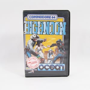 Puede incluir: Cartucho de videojuego Commodore 64 para "High Noon" con una ilustración de temática western. La caja del juego tiene un marco negro con el título del juego en azul y blanco, y el logotipo de Ocean Software.