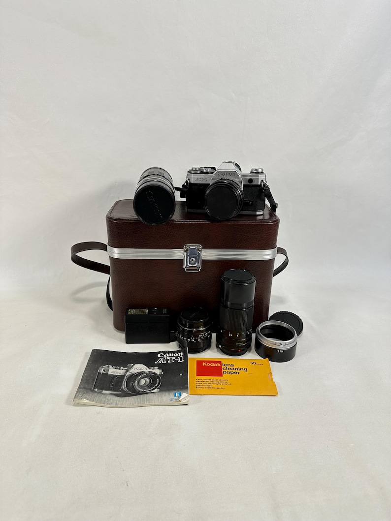 May include: A vintage silver Canon AE-1 camera with a brown leather carrying case, three lenses, a flash, and a Kodak lens cleaning paper. The camera has a black lens cap and a silver Canon logo.