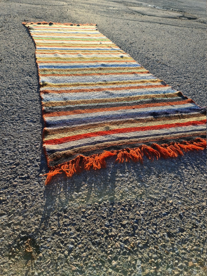 May include: A long, colorful striped rug with orange fringe. The rug is made of woven fabric and has a variety of colors, including red, orange, yellow, green, blue, and brown.