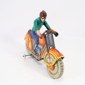 May include: Vintage toy motorcycle with a rider. The motorcycle is primarily orange with yellow and black accents. The rider is wearing a green shirt, blue pants, and a brown helmet. The toy has a pull-back wheel.