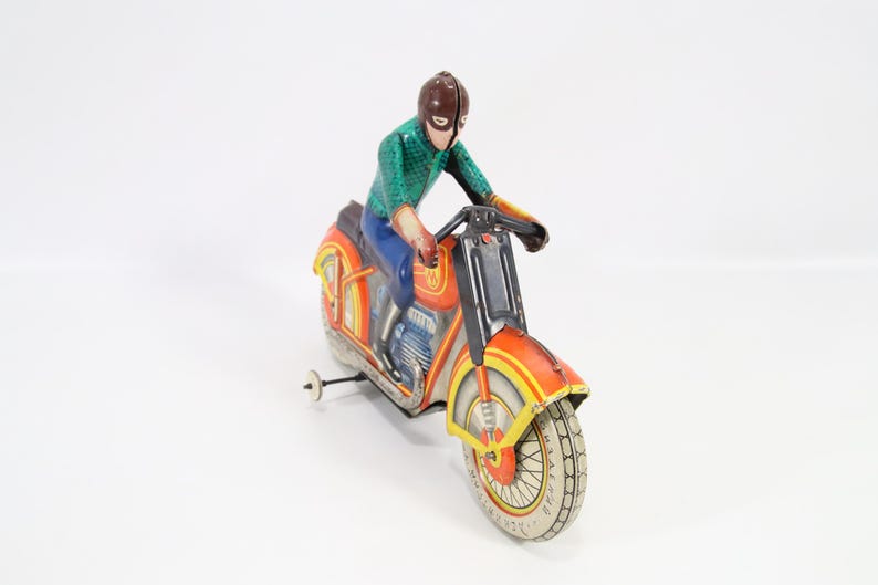 May include: Vintage toy motorcycle with a rider. The motorcycle is primarily orange with yellow and black accents. The rider is wearing a green shirt, blue pants, and a brown helmet. The toy has a pull-back wheel.